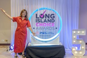 Woman in a red dress posing with arms outstretched beside the Long Island Choice Awards backdrop featuring a glowing circular sign and large text.