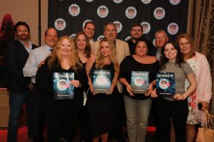 Group of people posing together, holding WINNER plaques at a Long Island Choice Awards backdrop with blue circular logos on a dark curtain behind them.