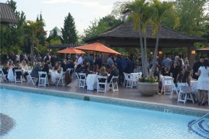 Guests mingling at a poolside outdoor reception with white-clothed tables under orange umbrellas and a pavilion in the background.