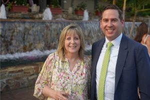 Smiling woman in a floral blouse and a man in a navy suit posing together outdoors in front of a stone fountain.