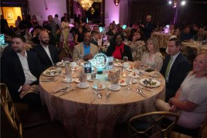 Group of ten people in formal attire seated around a round banquet table at a gala, with a glowing centerpiece and table settings visible around them, in a dimly lit event hall.