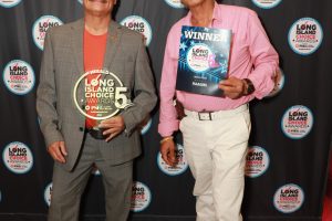 Two men pose at a Long Island Choice Awards backdrop; left man in a gray suit holds a circular award, right man in a pink shirt holds a 'WINNER' magazine.