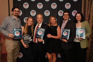 Six adults stand together at an awards event posing for a group photo; some hold 'WINNER' plaques and one holds a circular Long Island Choice Awards trophy, with a backdrop covered in repeating Long Island Choice Awards logos.