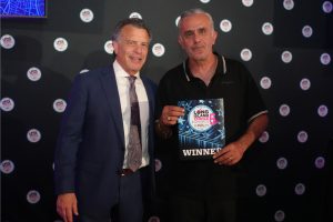 Two men posing at an awards event; one in a blue suit and patterned tie, the other in a black polo holding a 'WINNER' certificate with star graphics.
