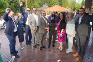 Group of professionally dressed adults posing together outdoors at a celebratory event, some raising fists and holding drinks near a brick patio and orange umbrella.