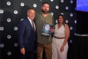 Three people pose for a photo at an awards event; center man holds a plaque that reads WINNER, flanked by a suited man and a woman in white, against a backdrop with circular logos.