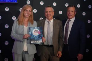 Three adults in business attire smile for a photo at an awards event; the woman on the left holds a 'WINNER' certificate for Long Island Choice Awards.