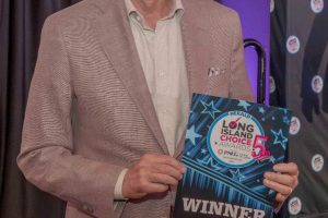 Smiling man in a beige blazer posing with a WINNER sign at an awards event.