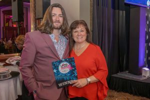 Two people smile for a photo at an awards event; man in a mauve suit and patterned shirt beside a woman in red, who holds a 'WINNER' certificate from Long Island Choice Awards.