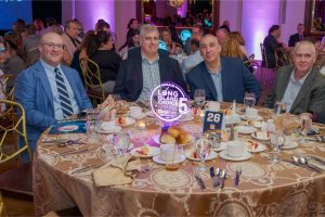 Four men in suits sit at a round banquet table during an awards dinner, with a neon 'Long Island Choice Awards' centerpiece.