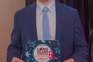 Young man in a blue suit and light blue tie holding a winner certificate at an awards event, smiling at the camera.