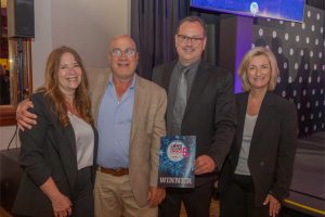 Four adults in business attire pose together at an awards ceremony; the man second from the right holds a blue 'WINNER' plaque.