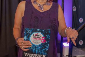 Smiling woman in a purple dress holds a blue Long Island Choice Awards 'WINNER' badge on stage.