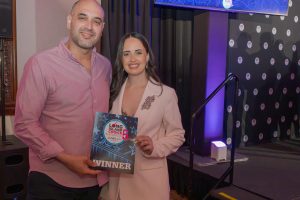 Two casually dressed people pose together, holding a winner certificate at an awards event.