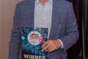 Man in a blue blazer holds a WINNER award plaque at an event, smiling at the camera in front of a dark curtain.