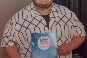Person wearing glasses and a geometric shirt holds a blue 'WINNER' plaque at an awards event.