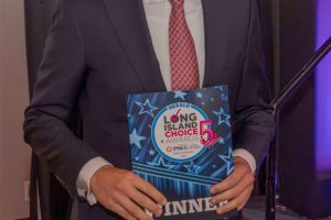 Man in a dark suit and red tie smiles while holding a Long Island Choice Awards program at a formal event.