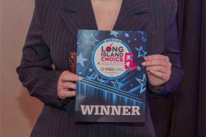 Woman in a dark pinstripe suit holds a blue award plaque reading WINNER at a ceremony photographer-style shot with a dark curtain backdrop.