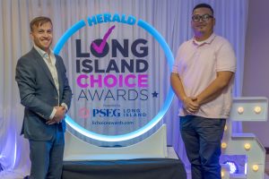 Two men pose beside a glowing circular sign that reads 'Long Island Choice Awards' on a branded backdrop.aced by a man in a suit and another in a light pink polo shirt.