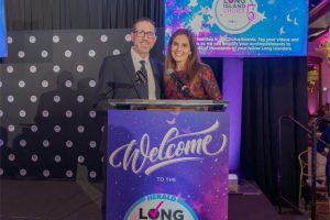 Two smiling hosts stand at a podium on a stage during an awards ceremony, microphones in front of them and a purple welcome banner on the podium.