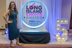 Smiling woman in a teal dress and tan heels stands beside an illuminated circular frame with the 'Long Island Choice Awards' backdrop behind her.