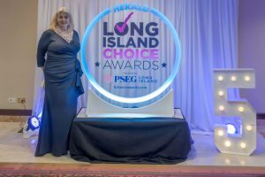 Woman in a navy evening gown posing beside a glowing circular sign that reads Herald Long Island Choice Awards, at a ceremonial backdrop.