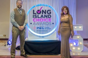 Man in a tan vest and tie and a woman in a shimmering gown pose on a stage beside a blue circular sign reading 'Long Island Choice Awards'.