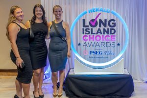 Three women in black dresses pose together at an awards ceremony next to a glowing Long Island Choice Awards sign.