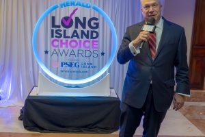 Man in a navy suit and red striped tie speaks into a microphone at the Long Island Choice Awards, standing beside a circular blue-lit sign labeled 'Herald Long Island Choice Awards'