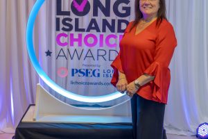 Woman in red blouse standing beside a circular glow sign reading 'Long Island Choice Awards' with a draped backdrop, smiling at the camera.