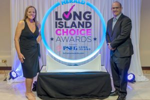 Woman in a black dress and a man in a suit stand on either side of a glowing circular award sign reading 'HERALD LONG ISLAND CHOICE AWARDS' with 'Presented by PSEG Long Island' in the center.