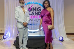 Two adults pose on a stage at an awards backdrop, a man in a light gray pinstripe suit and a woman in a magenta satin dress.