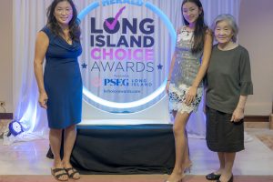Three women pose in front of a Long Island Choice Awards backdrop with a blue neon circular frame on stage, smiling at the camera.