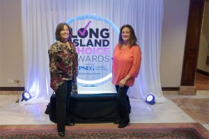 Two women pose and smile in front of a circular lit sign that reads 'Herald Long Island Choice Awards' with white curtains behind them.