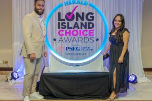 Two people pose beside a glowing circular sign that reads 'Herald Long Island Choice Awards' with a white draped backdrop.