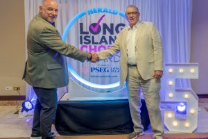 Two men in blazers shake hands on a stage in front of a neon-lit circular sign reading 'Herald Long Island Choice' backdrop.