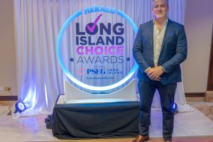 Man in a blue suit posing next to a circular blue neon sign reading 'Long Island Choice Awards' with a white draped backdrop behind him
