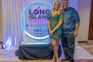 Couple posing at the Long Island Choice Awards backdrop with a neon circular logo behind them, smiling for the camera.
