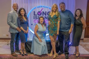 Six adults pose for a group photo at an indoor event, with a glowing circular backdrop reading 'Herald' and 'Long Island' behind them—the two men and four women in smart-casual attire.