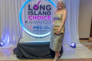 Woman in a gold strapless gown posing with a tan clutch at the Long Island Choice Awards backdrop circle.Backdrop reads 'HERALD LONG ISLAND CHOICE AWARDS' with neon blue circle around it.