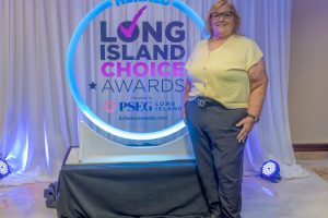 Woman posing next to a backlit circular sign that reads 'Long Island Choice Awards' with 'Herald' branding, wearing a pale yellow top and dark pants. Staffed event backdrop with blue lighting and curtains in the background.
