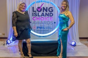 Two women in formal gowns pose at an awards backdrop with a lit circular sign that reads Herald Long Island Choice Awards.