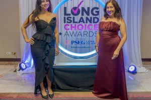 Two women in formal gowns pose on a stage in front of a Long Island Choice Awards backdrop with a glowing circle design and curtains behind them.