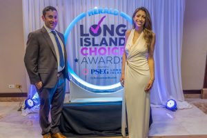 Man in a gray suit and blue tie and a woman in a light-colored evening gown pose beside a circular illuminated sign reading 'Long Island Choice Awards' at a backdrop with blue lighting and white drapes.