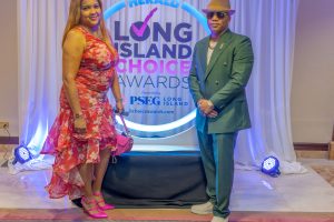 Two people pose on a red-carpet stage in front of a Long Island Choice Awards backdrop; woman in a pink floral dress and pink heels, man in a green suit and hat with sunglasses.