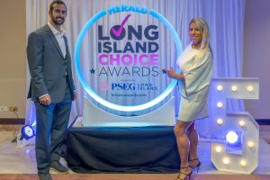 Two formally dressed individuals pose beside a glowing circular backdrop that reads 'Herald Long Island Choice Awards' at an awards event.
