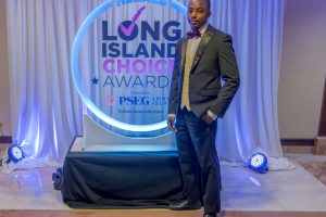 Man in a black tuxedo and purple bow tie posing beside a glowing 'Long Island Choice Awards' backdrop at a formal event.