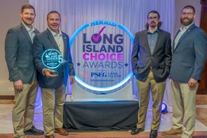 Four men in business attire stand in a row at an awards event beside a neon circular display reading 'Herald Long Island Choice Awards' with a trophy held by the second man from the left.