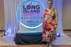 Woman in a red and pink floral dress stands beside a glowing circular 'Long Island Choice Awards' backdrop at an awards event.