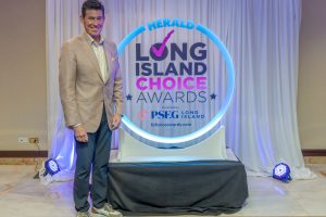 Smiling man in a beige blazer posing beside a glowing circular sign that reads Herald Long Island Choice Awards.